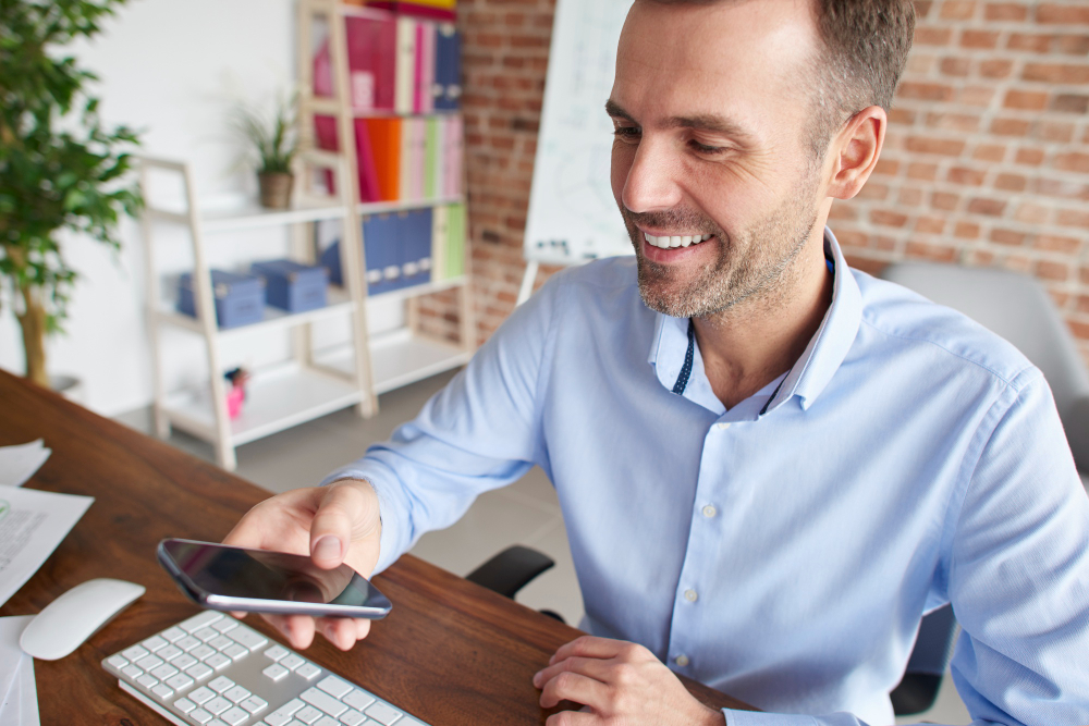 Homem branco olhando para tela do celular com expressão sorridente. Trata-se de um gestor selecionando os benefícios que estão em alta para a sua empresa